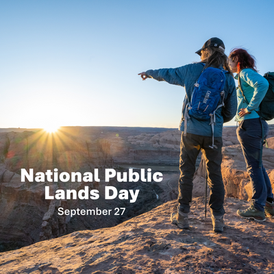 A banner for National Public Lands Day, a couple stand on a desert cliff pointing to the sun over the horizon