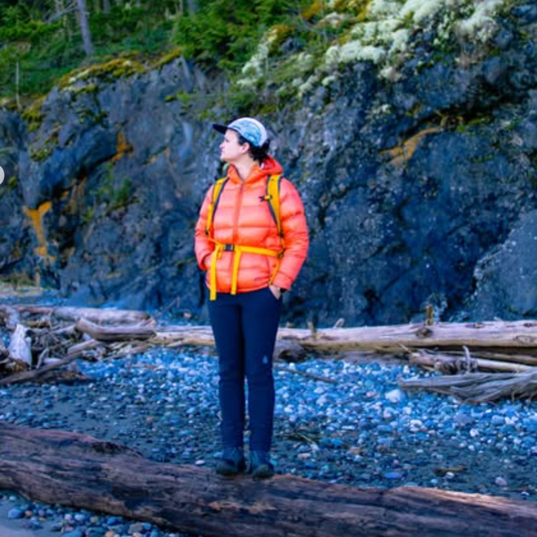 Person wearing an orange jacket standing on a rocky beach with a rocky cliff in the background