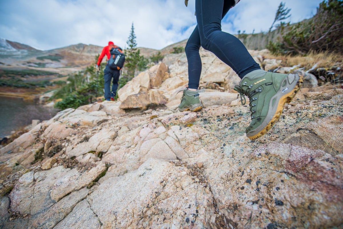 Two hikers walking along a rocky ridgeline. The one in focus is wearing sage green LOWA hiking boots and blue leggings. Another hiker in the distance is wearing red. 