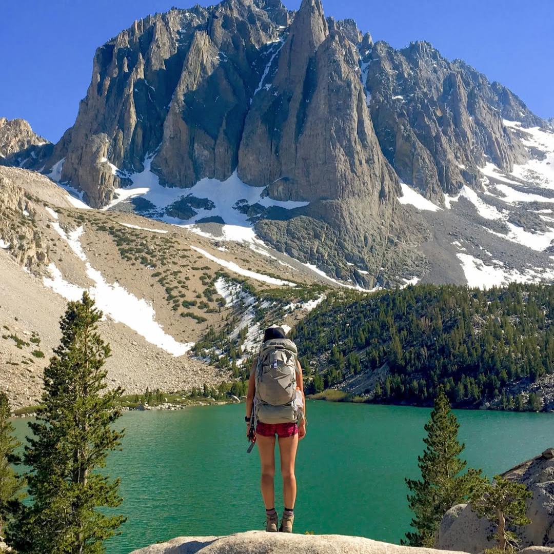 A person wearing a backpack and hiking boots faces a blue alpine mountain lake